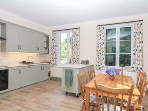 A modern kitchen with light-colored cabinets, a central island, and a dining area with wooden chairs and a table. - Lukesland Wing (Ivybridge)