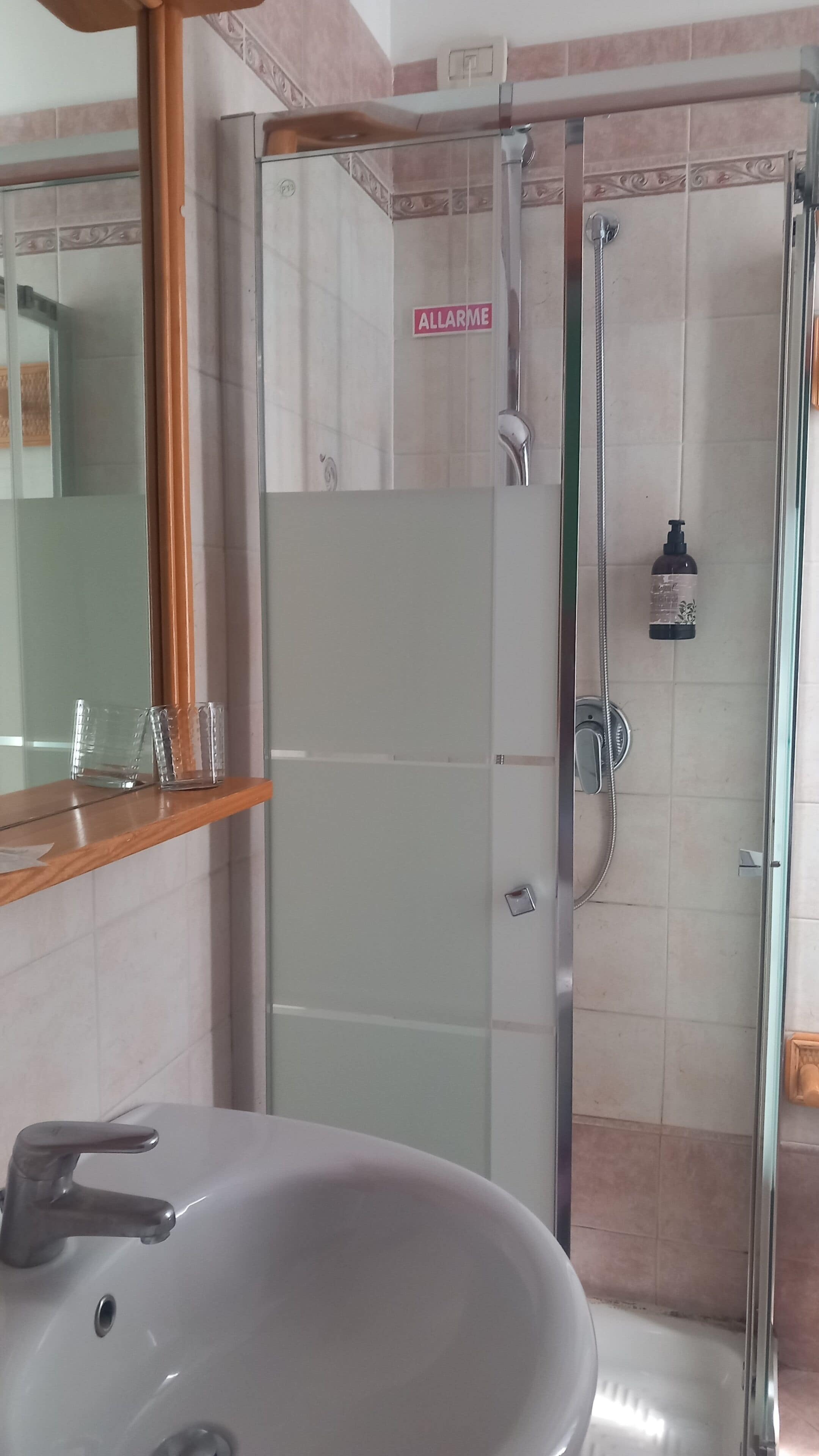 A bathroom with a glass shower enclosure, a sink with a mirror above it, and a sign that reads 'ALLARME'.