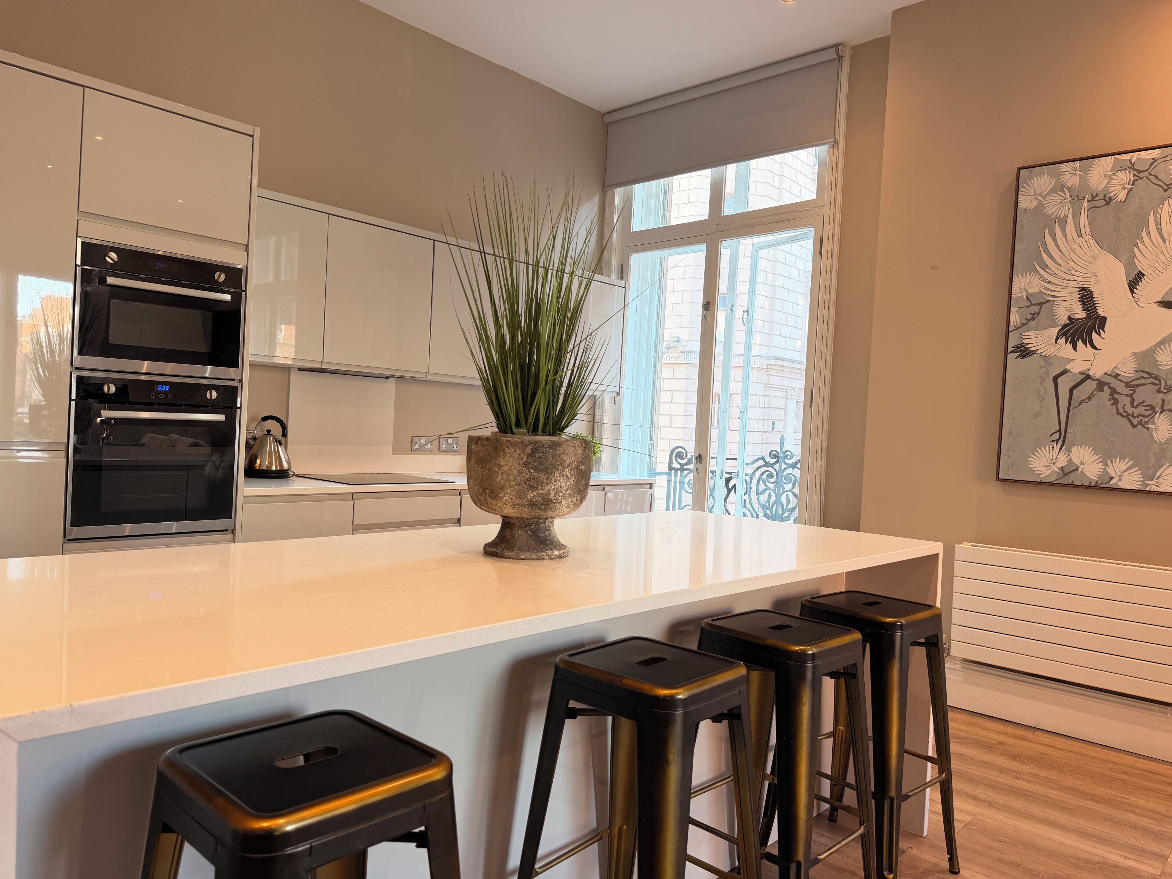 A modern kitchen with built-in ovens, a central island with stools, and a large window with a view of buildings.