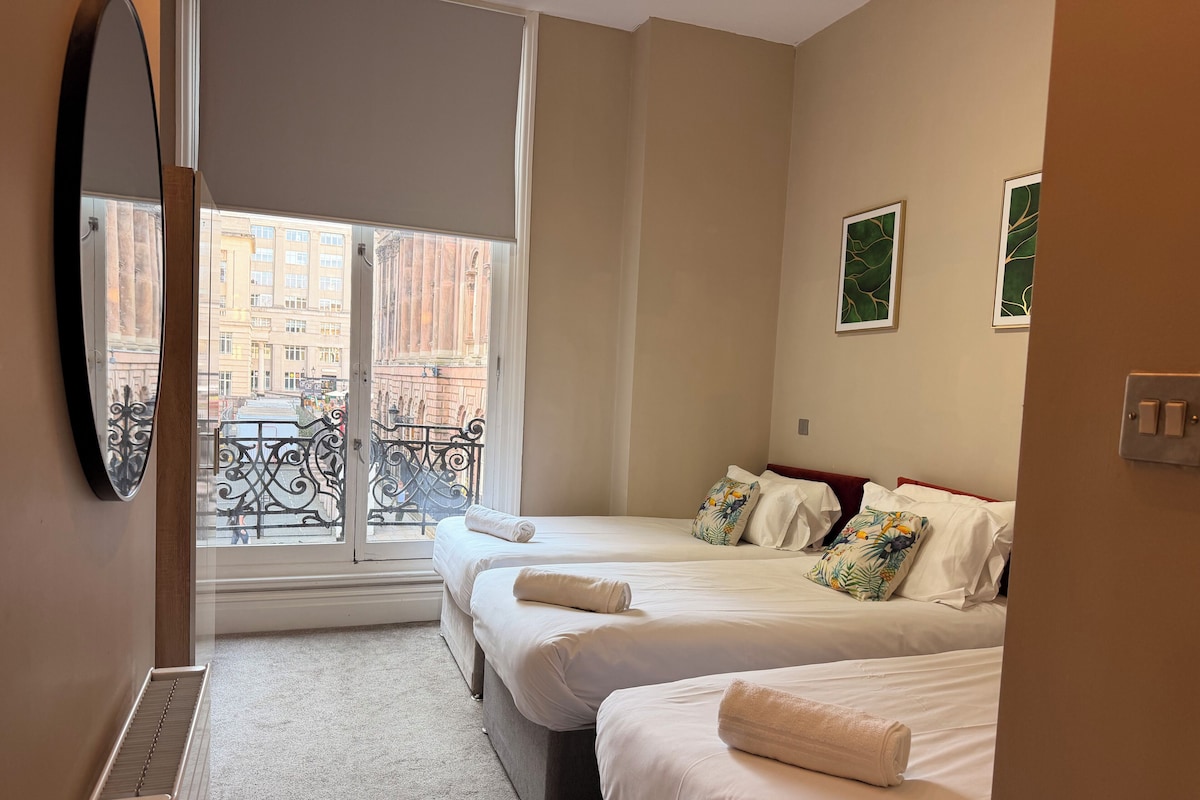 Una camera d'albergo con due letti, un'ampia finestra vista edifici, un termosifone e una porta.