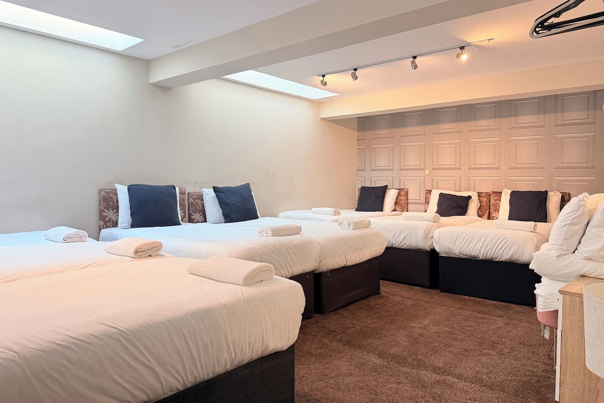 Camera d'albergo con quattro letti allineati, ognuno con un asciugamano piegato con cura. Ampia finestra che offre luce naturale, e l'ambiente presenta un design moderno con colori neutri.
