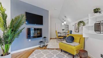 A modern living room with a flat-screen TV, a yellow sofa, a dining area, and a large potted plant.