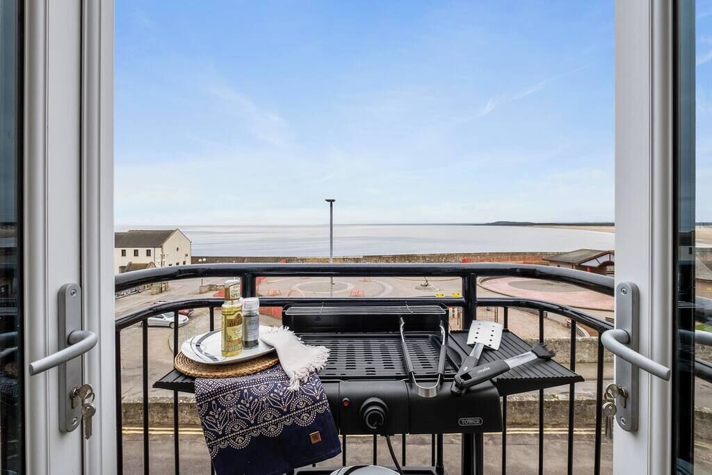 A balcony with a grill, a table with a bottle and a plate, and a view of a coastal road.