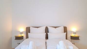Ein Doppelbett mit weißen Bettlaken und zwei Kissen, seitlich flankiert von Nachttischen mit Lampen, in einem Zimmer mit schlichten weißen Wänden.