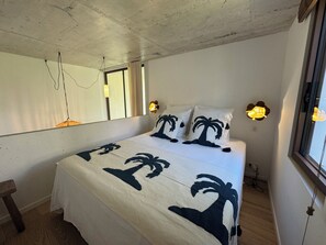 A bedroom with a bed, a bedside table, a mirror, and a window with curtains. - Architect-designed villa with a pool and a view of the Mediterranean – Sleeps 9 (Rayol-Canadel-sur-Mer)
