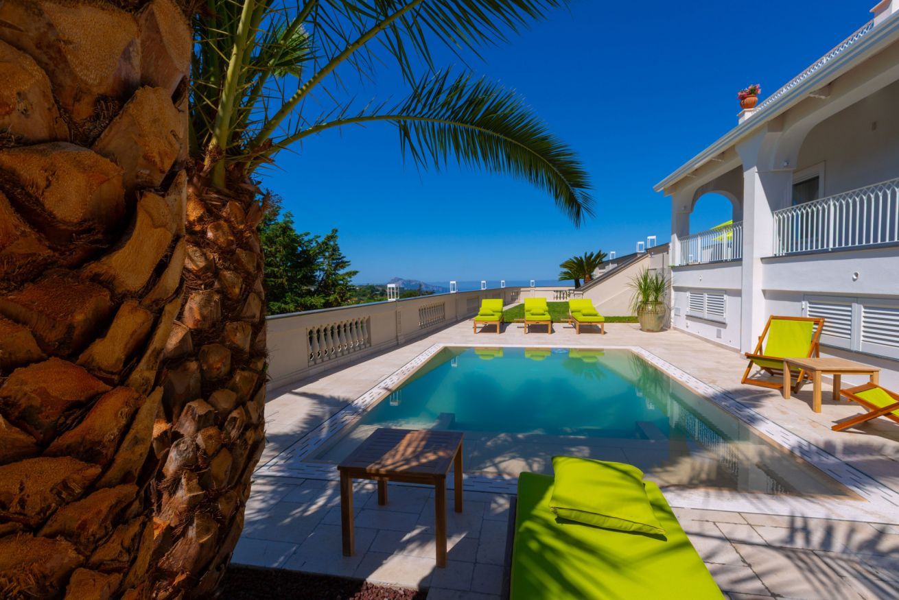 A pool area with lounge chairs, a palm tree, and a view of the sea.