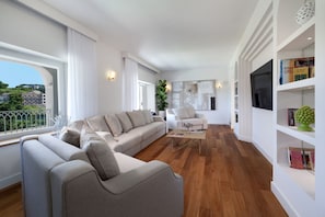 Uma sala de estar moderna com um sofá, poltronas e uma mesa de centro. Há uma estante com livros e objetos decorativos.