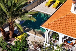 Uma área de piscina no terraço, com uma palmeira, cadeiras de descanso amarelas e um edifício branco com telhado de azulejos.