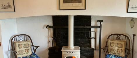 A cozy room with a white wood-burning stove, two chairs with patterned cushions, and framed pictures on the walls.