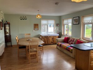 Ein gemütliches Wohnzimmer mit Esstisch aus Holz, einer Couch mit bunten Kissen und einer Küchenzeile mit Holzschränken.