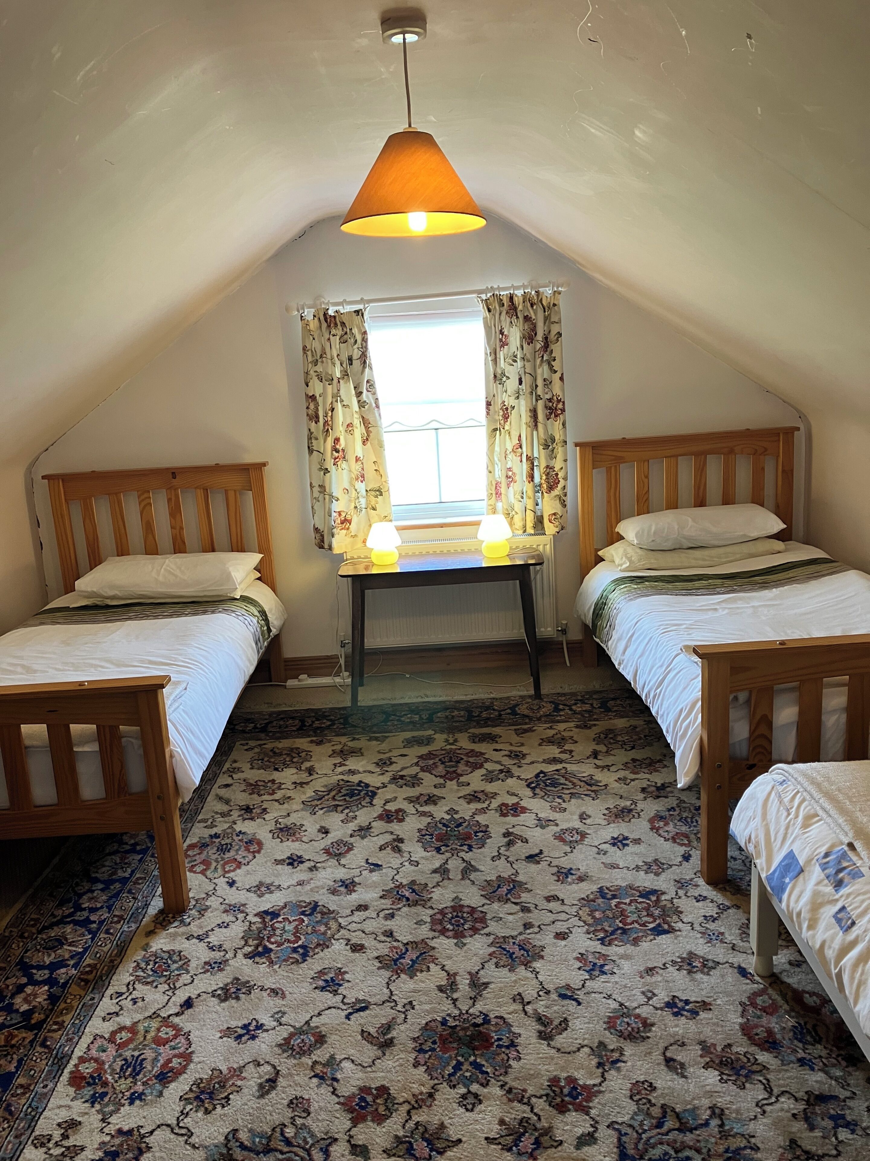 A room with two wooden beds, a table with lamps, a window with curtains, and a patterned carpet.
