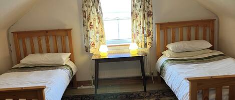 A room with two wooden beds, a table with lamps, a window with curtains, and a patterned carpet.