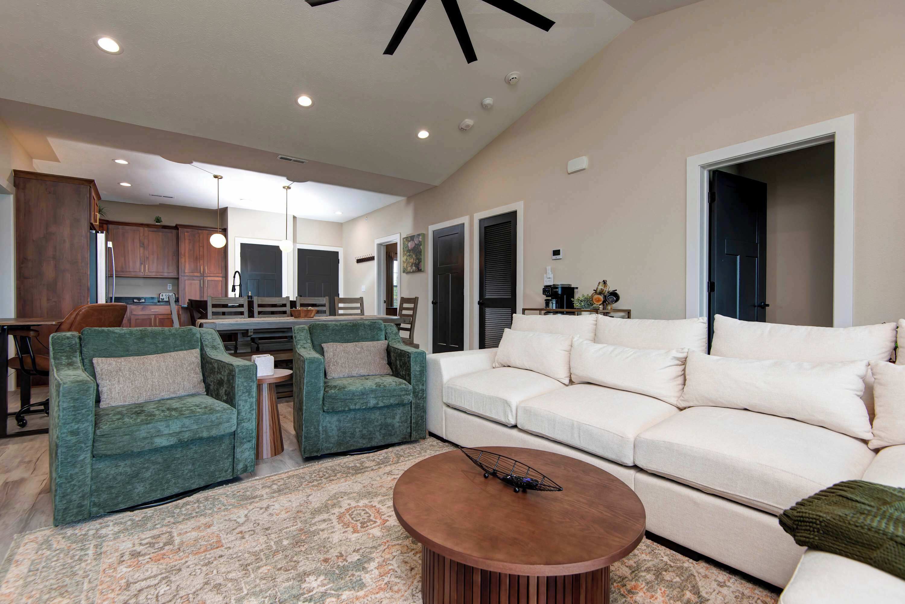 A spacious living room with a large white sofa, two green armchairs, a coffee table, and a kitchen in the background.