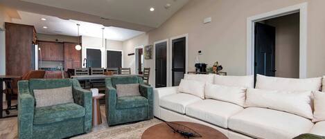A spacious living room with a large white sofa, two green armchairs, a coffee table, and a kitchen in the background.