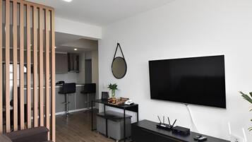 A modern living room with a flat-screen TV, a sofa, a coffee table, and a dining area with a kitchen in the background.