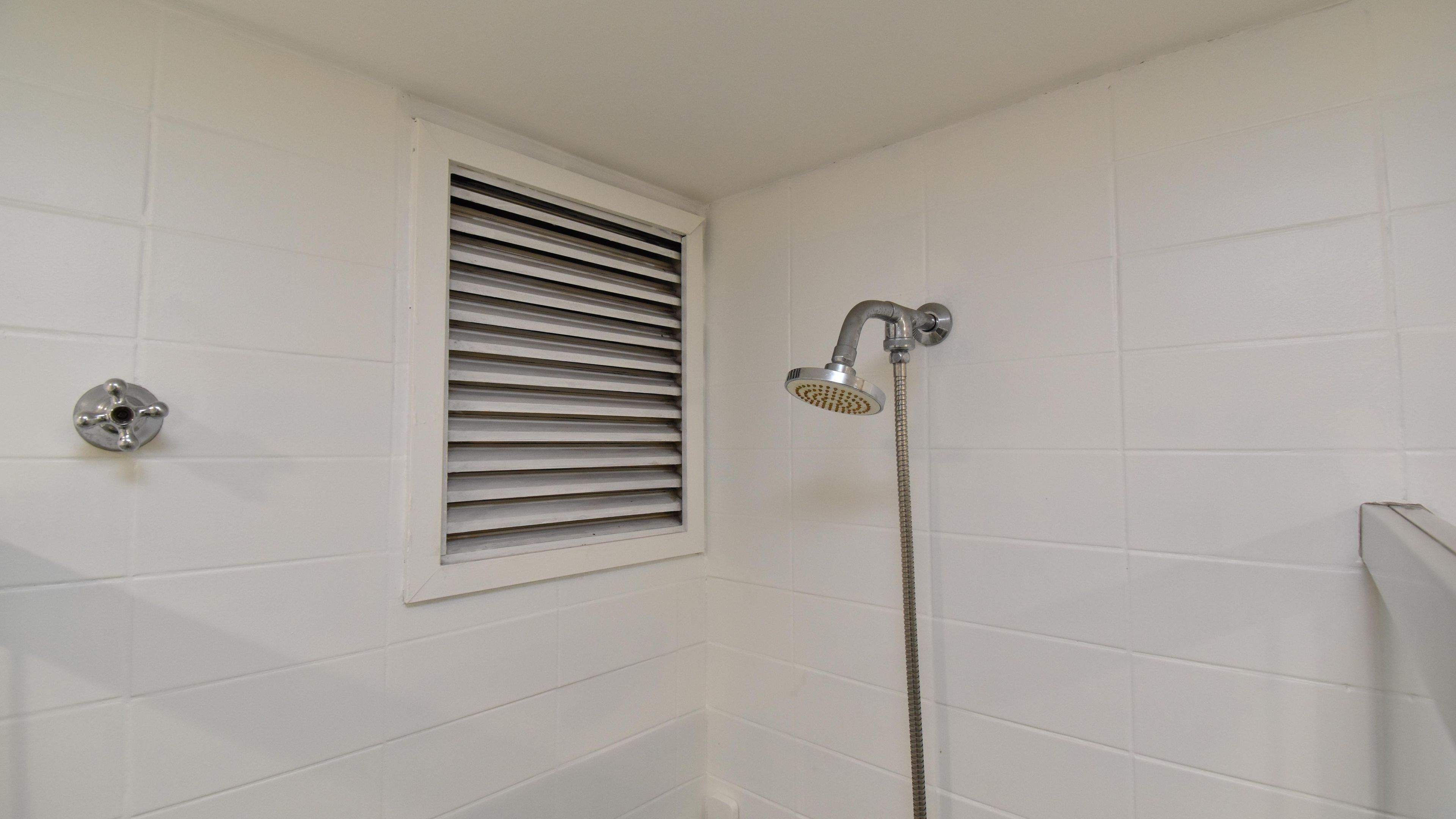 A bathroom with white tiled walls, a shower with a handheld showerhead, and a window with horizontal blinds.