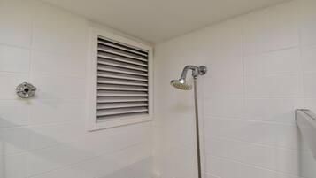 A bathroom with white tiled walls, a shower with a handheld showerhead, and a window with horizontal blinds.