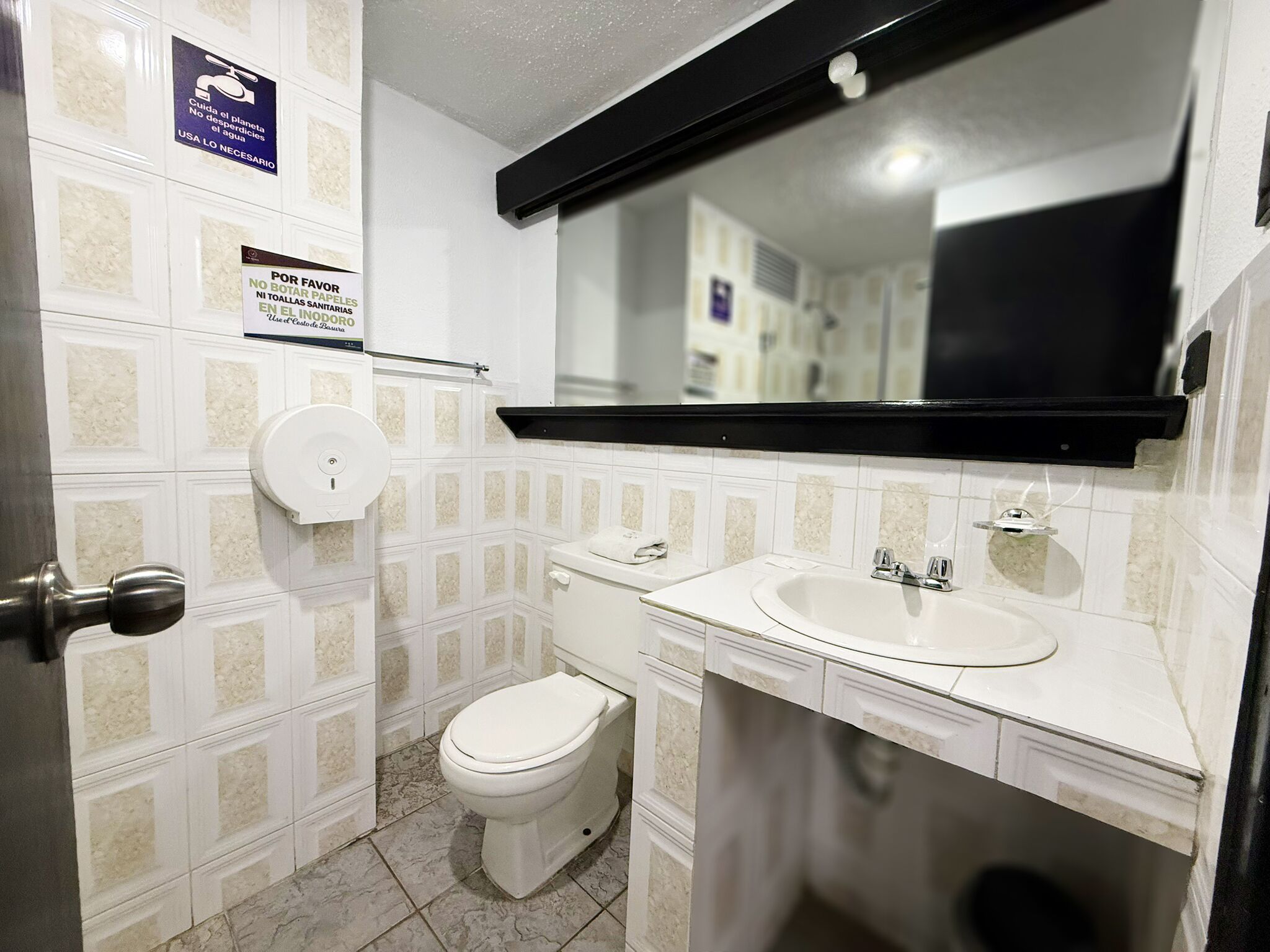 Un baño pequeño con inodoro, lavamanos y espejo. Hay carteles en la pared, uno de los cuales indica "Se requiere el uso de agua".