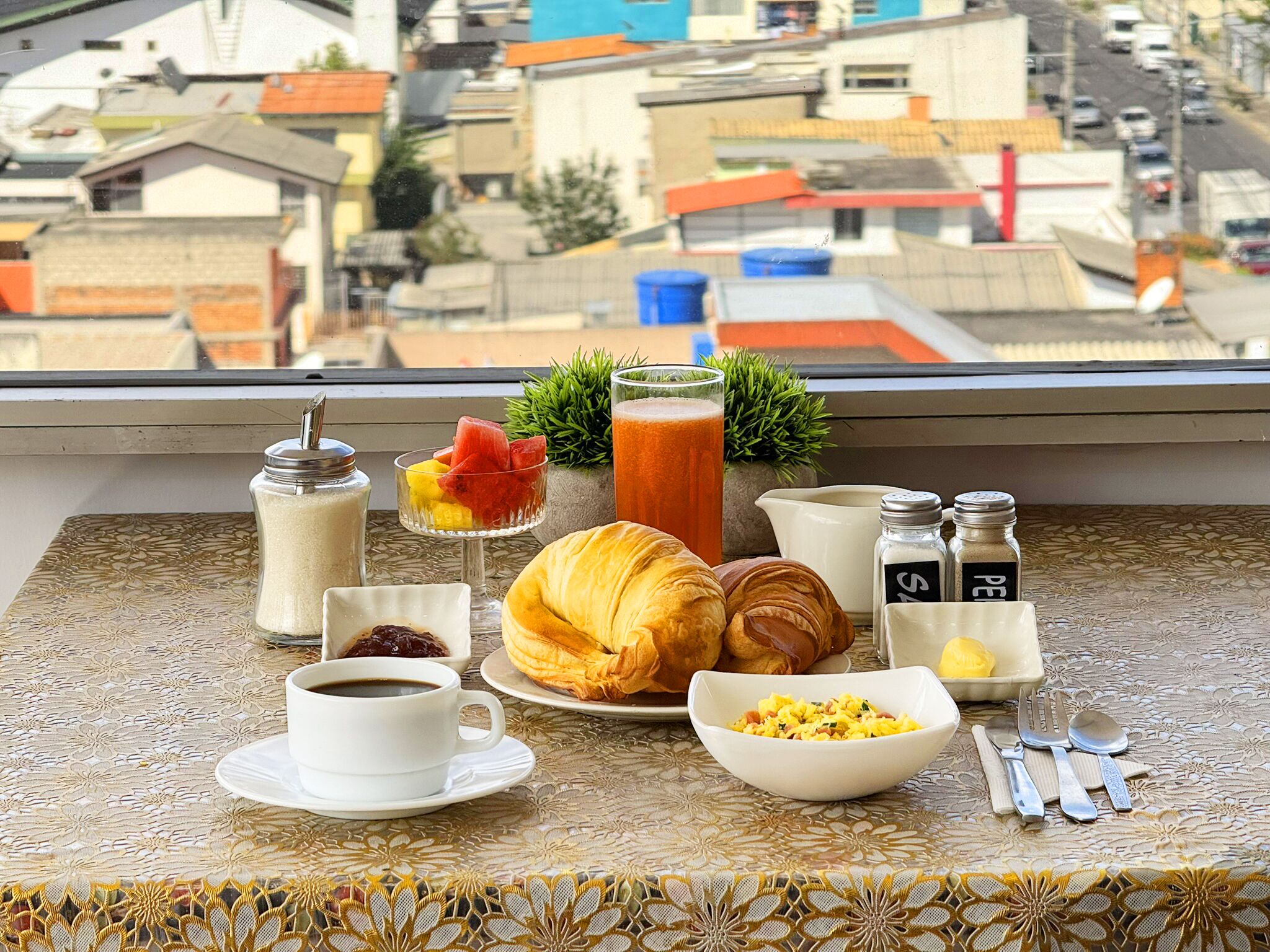 A breakfast setup with croissants, coffee, and fruit on a table by a window overlooking a cityscape.