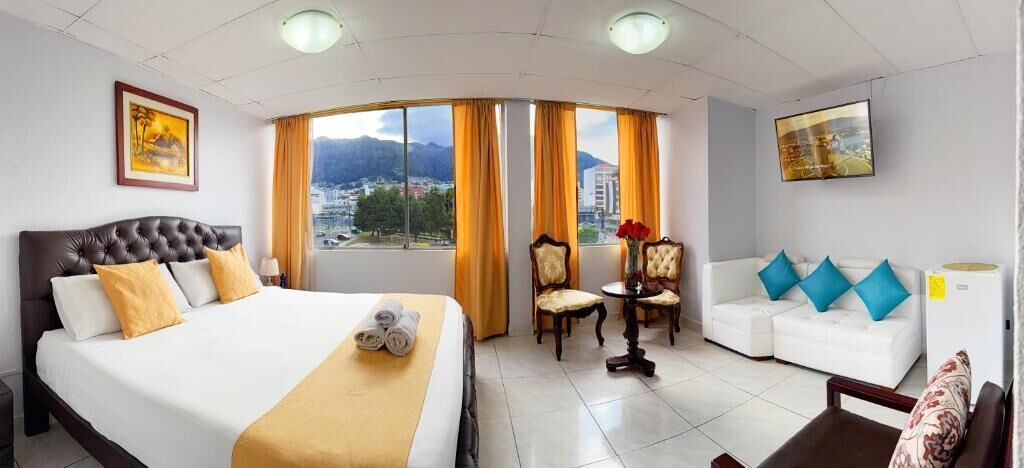 Habitación de hotel con una cama grande, un sofá, dos sillones, una mesita y vistas a la ciudad a través de amplias ventanas.
