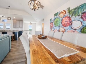 A modern kitchen with a central island, a dining area with a wooden table, and a living space with a sofa. - Seabatical-4x4, Semi-Oceanfront w/ Pool & Hot Tub (Carolla)