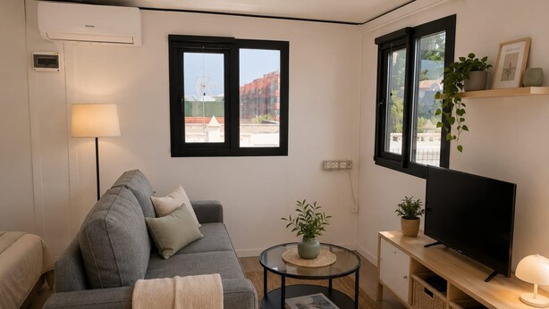 Un soggiorno moderno con un divano grigio, un tavolino rotondo con una pianta, una televisione su un mobile in legno e una finestra che si affaccia su edifici.