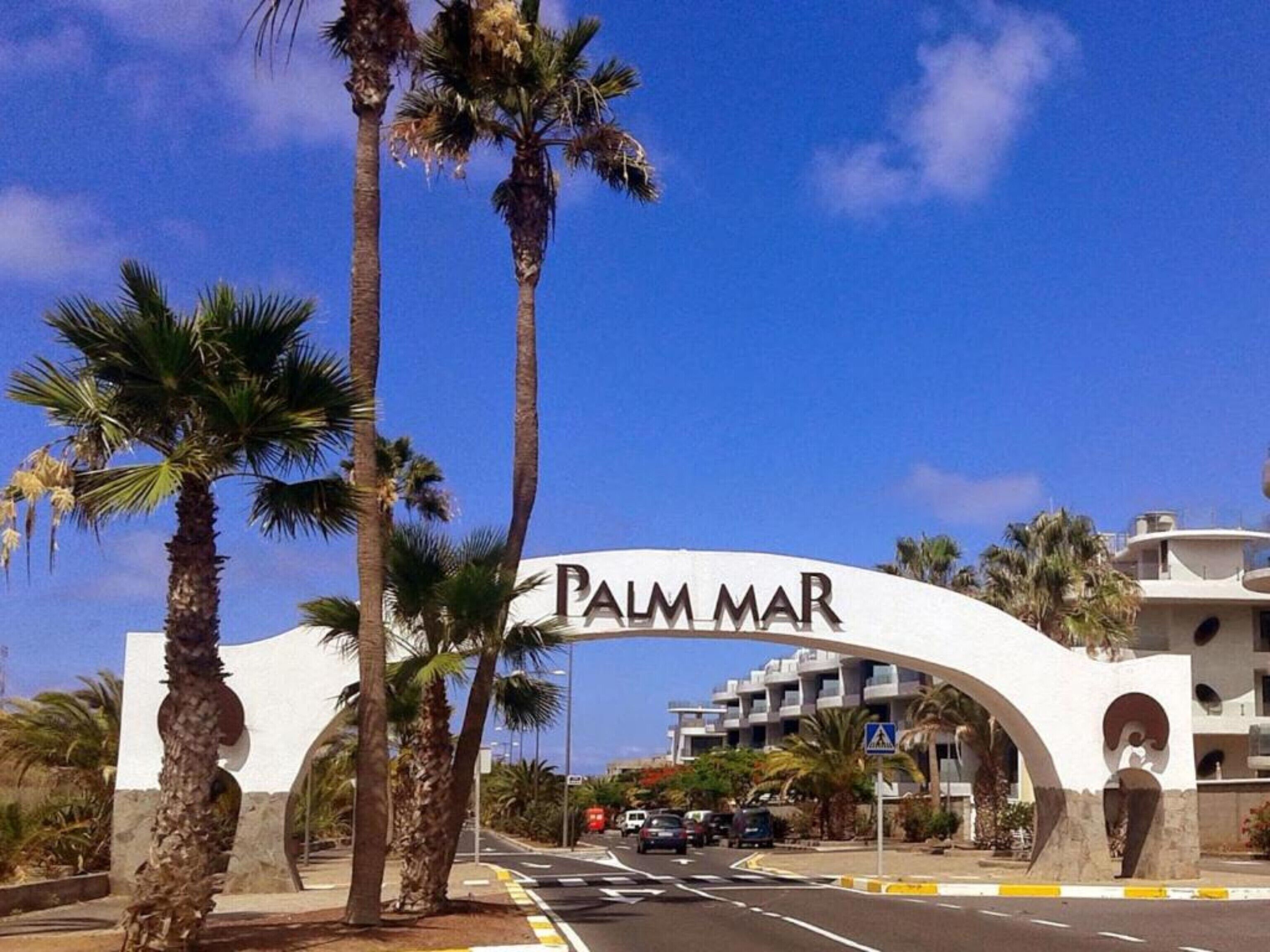 A street sign with 'PALM MAR' written on it, palm trees, and a building in the background.