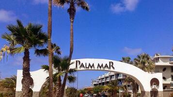 Una señal de calle con la leyenda 'PALM MAR', palmeras y un edificio al fondo.