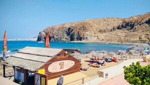 Una posizione fronte spiaggia con un edificio in legno, ombrelloni da spiaggia e un pendio collinare roccioso.