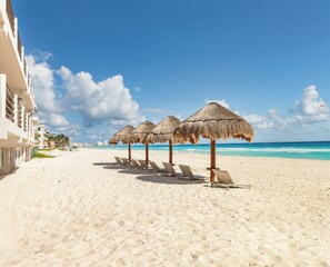 A beach with thatched umbrellas and sun loungers, a clear blue sky, and a building on the left. - Private Villa 3 in Cancún - Villas del Mar (Cancún)
