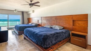 Un dormitorio con dos camas, cabecera de madera, ventilador de techo y un amplio ventanal con vista al océano.
