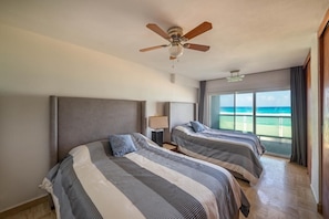 Une chambre d’hôtel avec deux lits, un ventilateur de plafond, une table de chevet, une lampe et une grande fenêtre donnant sur l’océan.