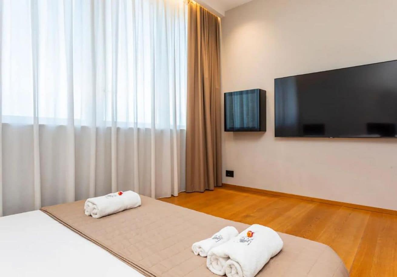 Ein Hotelzimmer mit Bett, zwei zusammengerollten Handtüchern, einem Flachbildfernseher und einem Fenster mit Vorhängen.