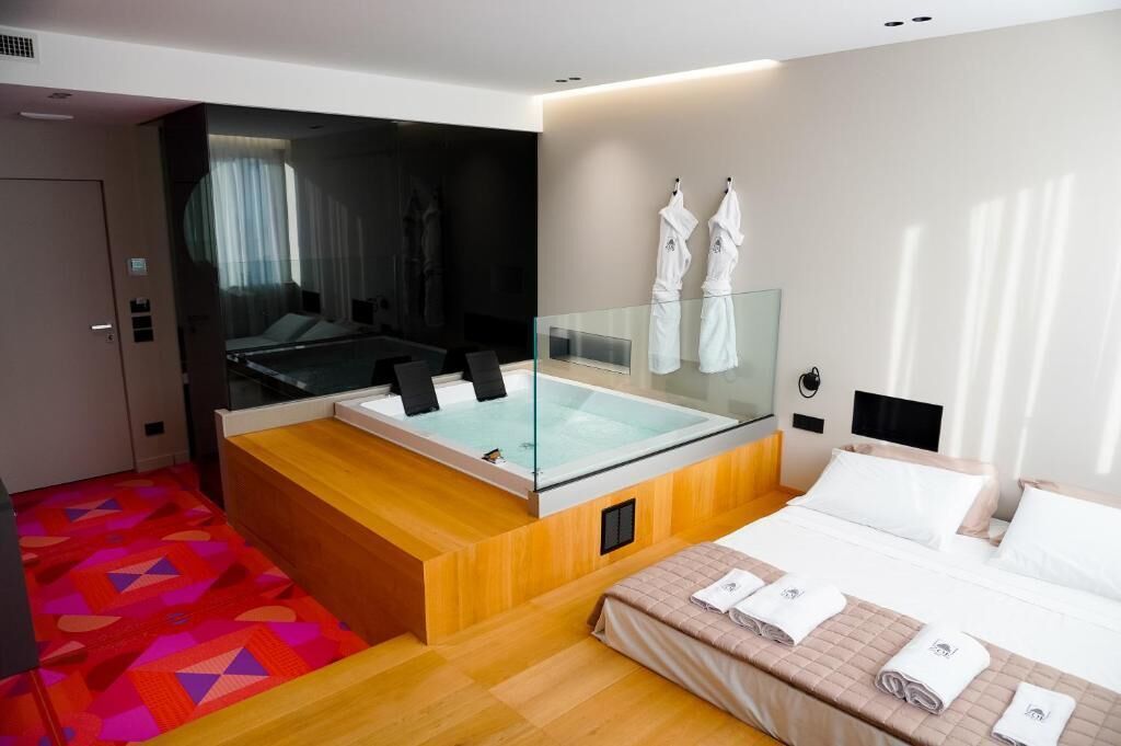 Una habitación de hotel moderna con un jacuzzi de vidrio, una cama con toallas ordenadas y una alfombra vibrante con un estampado geométrico.