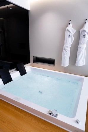 A large rectangular hot tub with a control panel, two white towels hanging on hooks, and a wooden floor. - Zoe luxury suites (Catania)