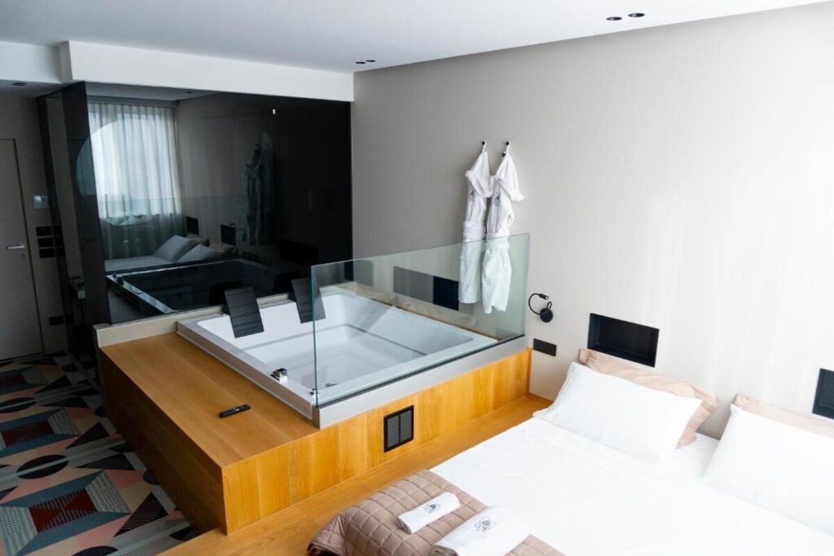 A modern hotel room with a glass-enclosed jacuzzi, a large bed, and a wooden bedside table.
