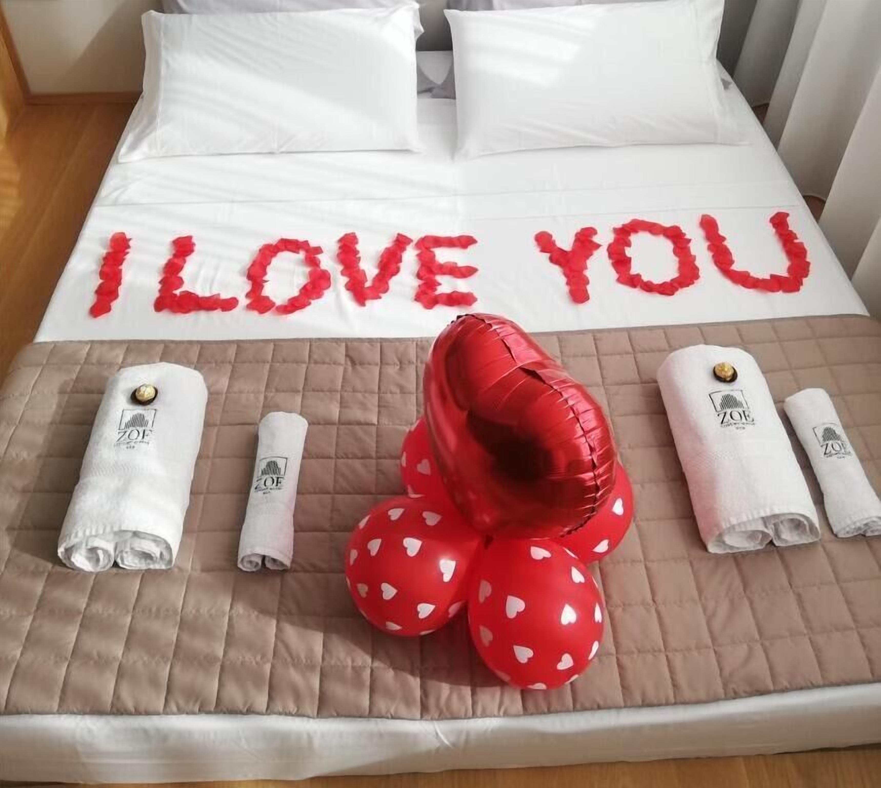 Un lit avec l’inscription « JE T’AIME » écrite en rouge, un coussin en forme de cœur également rouge, et des serviettes roulées ornées d’un logo.