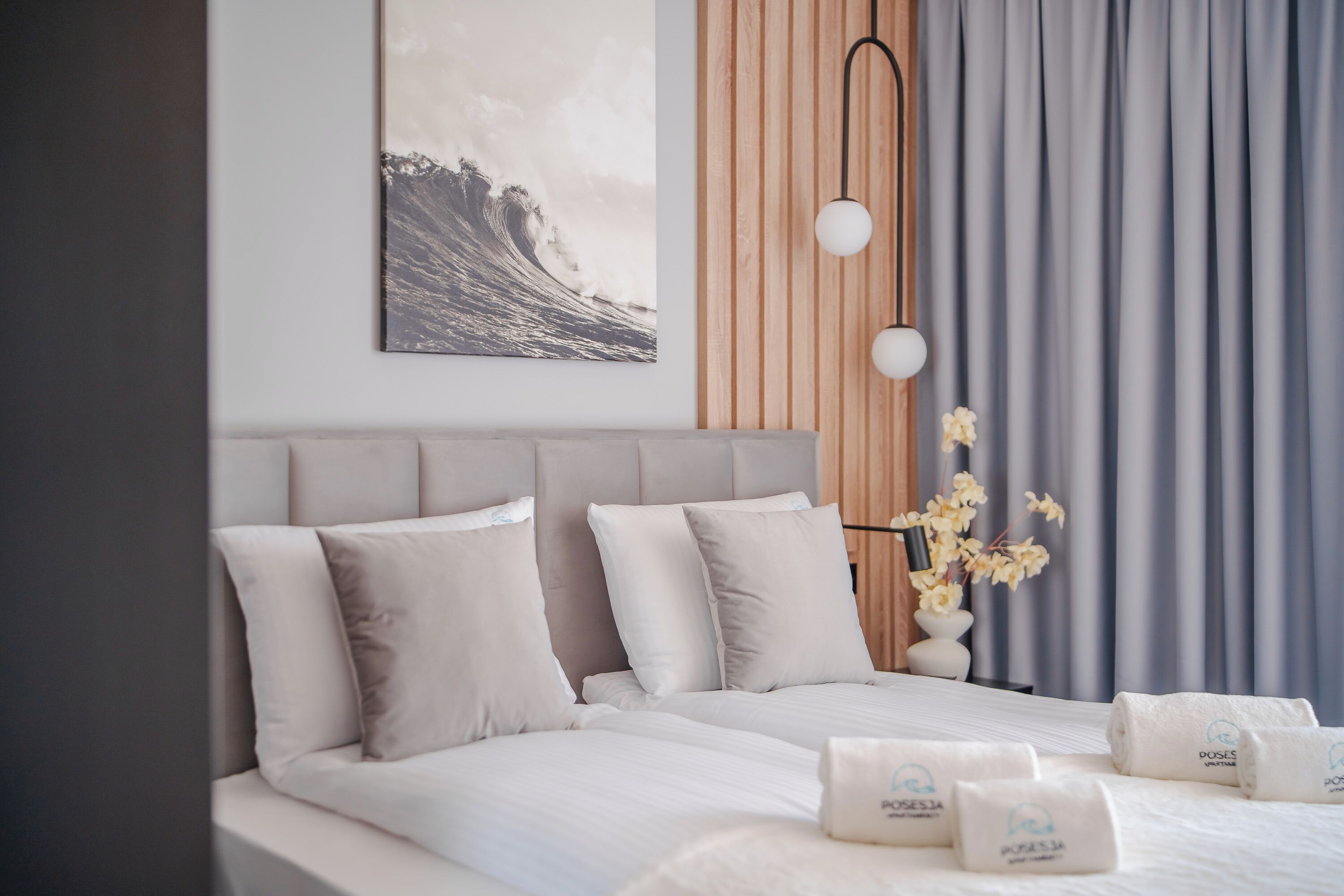 A modern hotel room with a large bed, a wooden headboard, a wall-mounted artwork, a bedside table with a lamp, and a vase with flowers.
