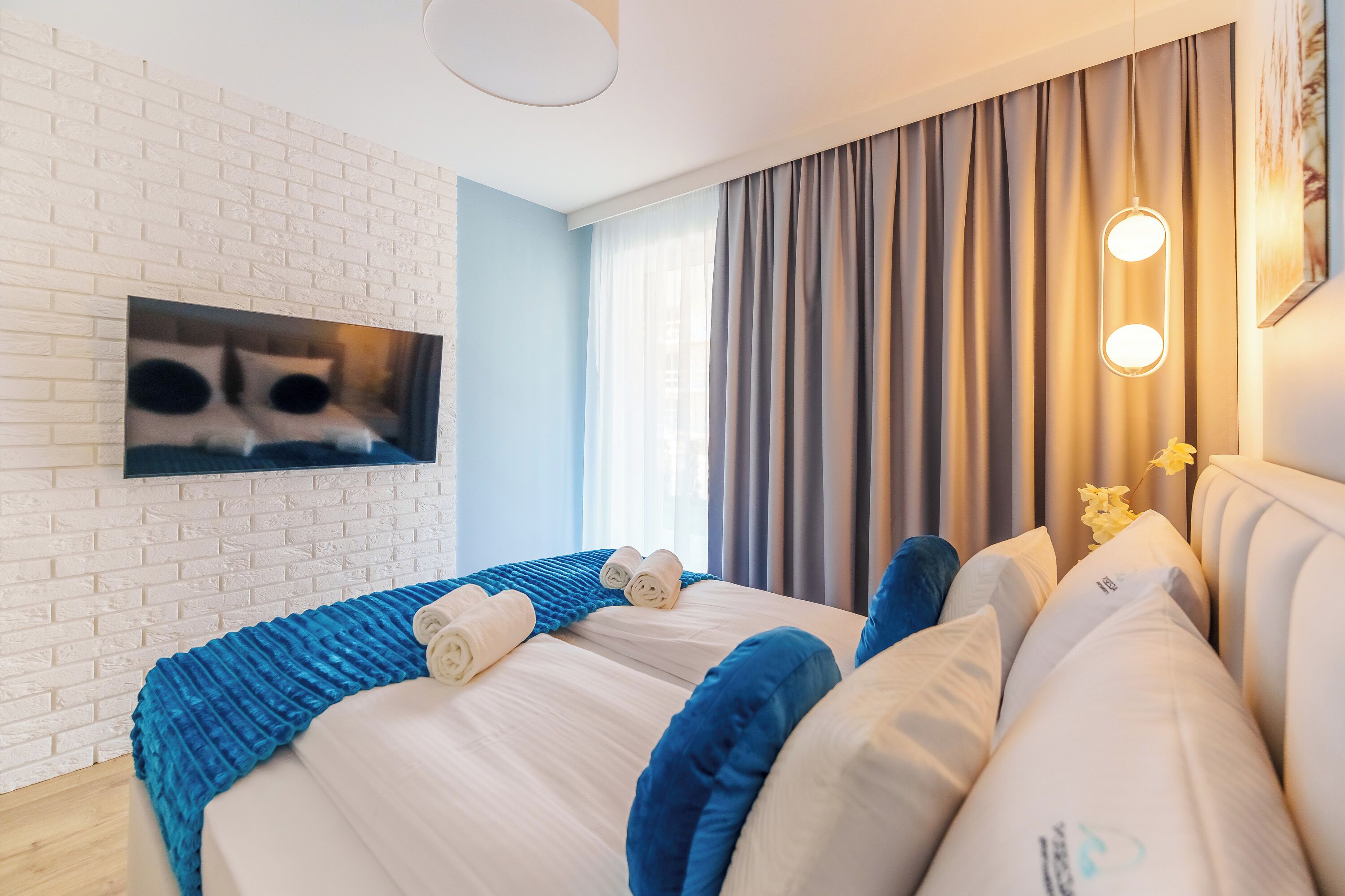 Camera d'albergo moderna con parete in mattoni, un letto grande con lenzuola bianche e azzurre, un televisore a schermo piatto montato a parete e un'ampia finestra con tende.
