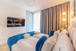 Una habitación de hotel moderna con pared de ladrillo, una cama grande con ropa de cama azul y blanca, un televisor de pantalla plana montado en la pared y un ventanal con cortinas.
