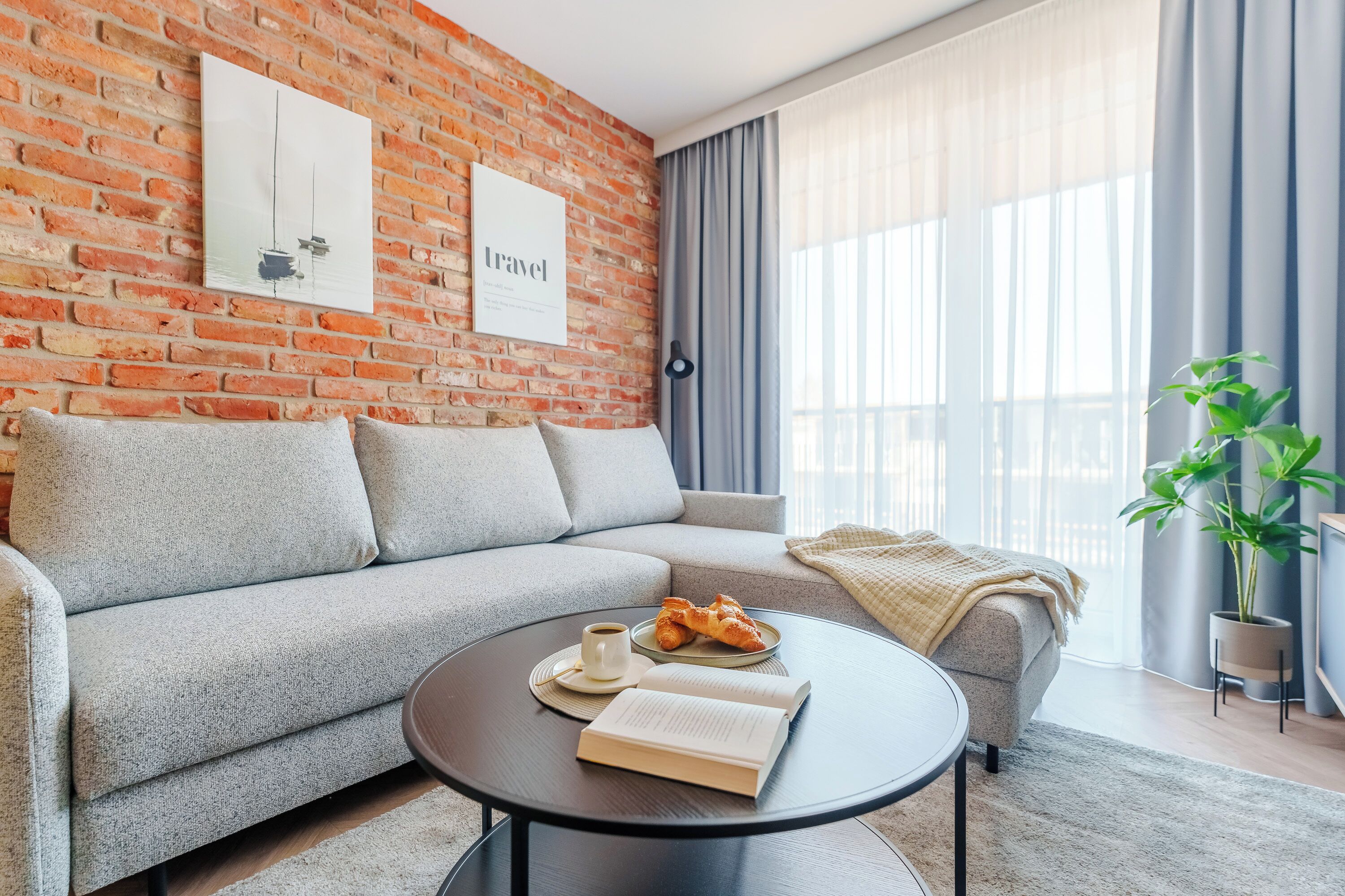 Ein modernes Wohnzimmer mit Backsteinwand, einer Couch, einem Couchtisch mit einem Buch und Croissants sowie einer Pflanze.