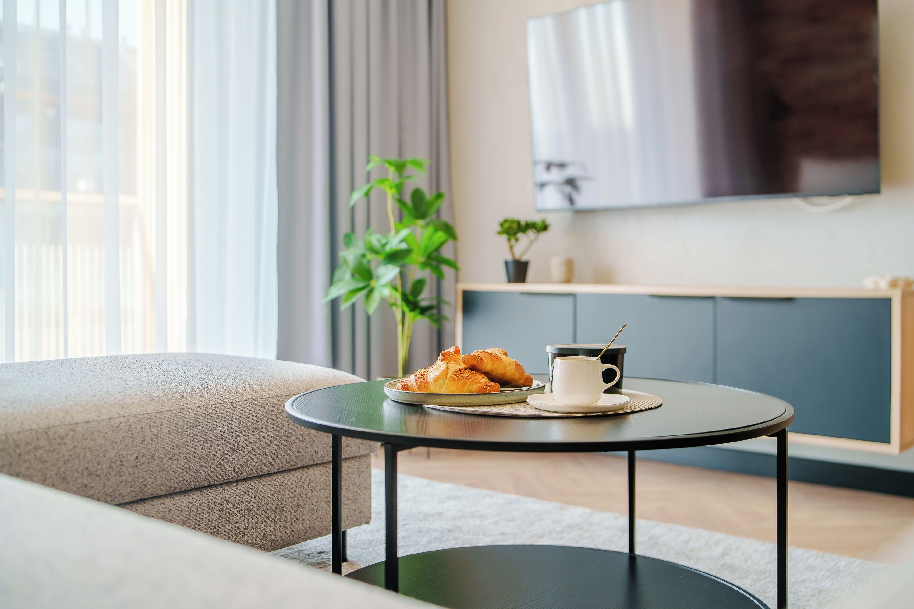 A modern living room with a round table, a sofa, a TV mounted on the wall, and a potted plant.