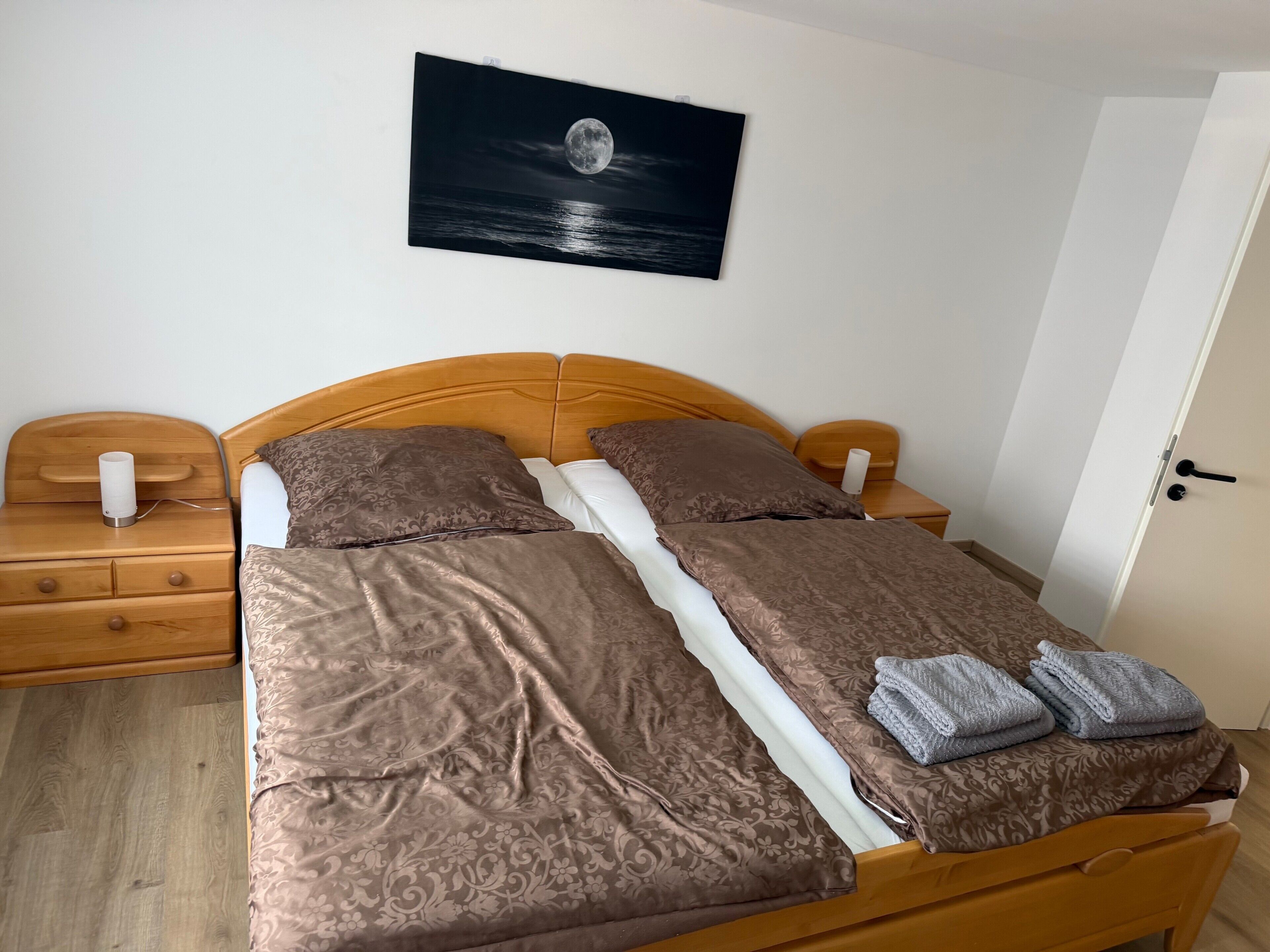 Un lit bien fait, avec une couvre-lit à motifs, un oreiller et une couverture. On y trouve une table de chevet en bois avec une lampe et un tiroir. Un tableau représentant une lune se reflétant sur l’eau est accroché au mur.