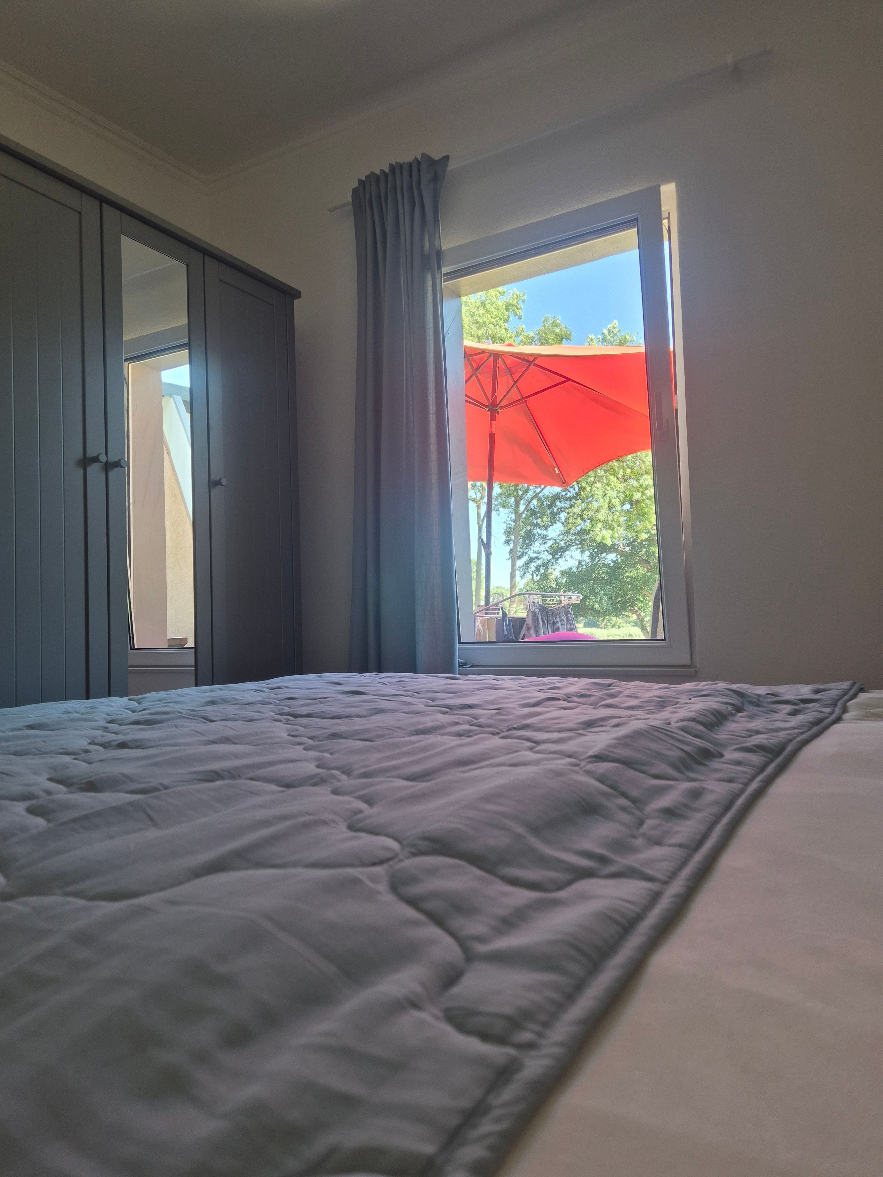 A bedroom with a bed, a wardrobe, and a window with a view of trees and an umbrella outside.