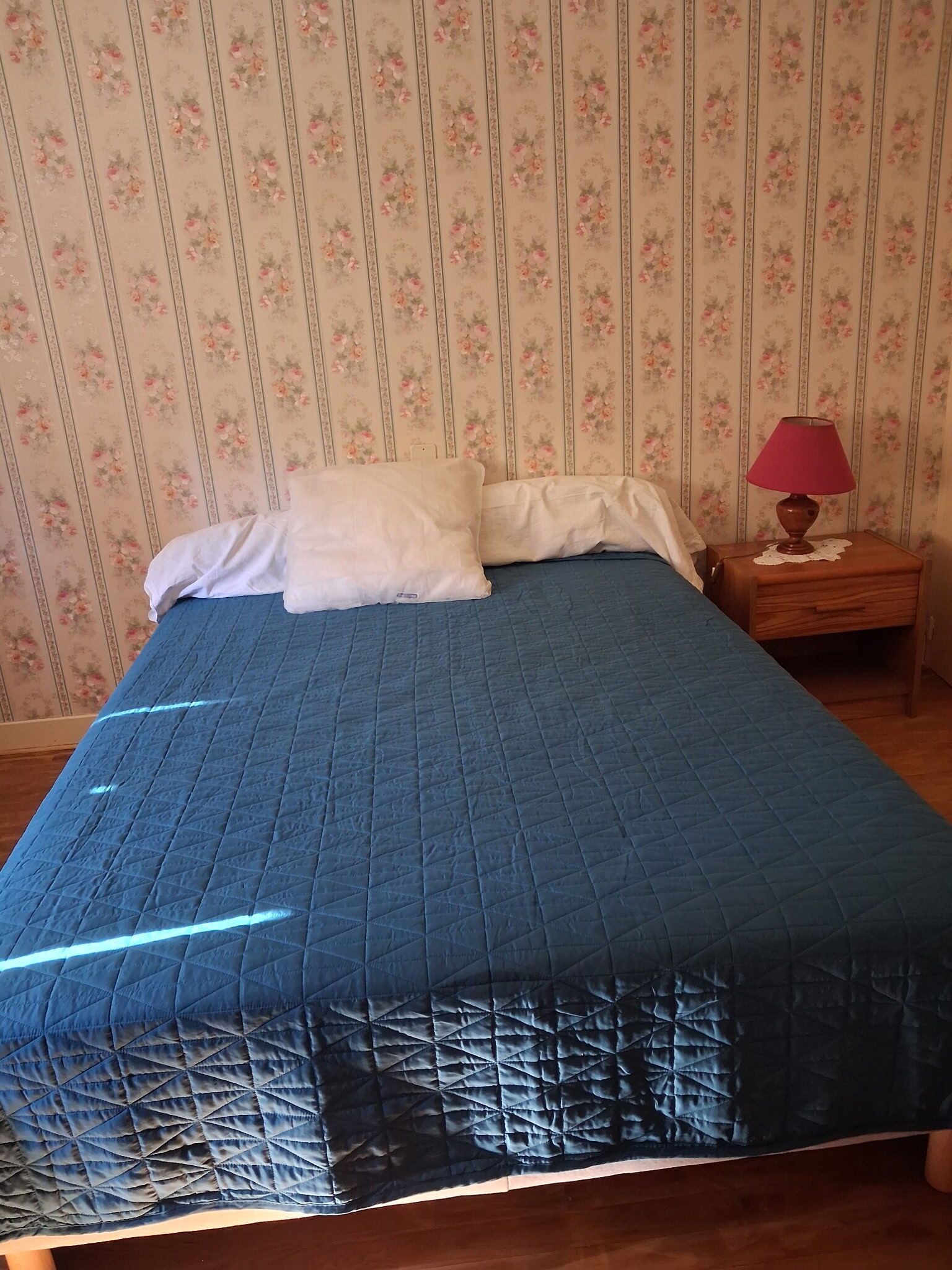 Un lit recouvert d’une couette bleue, une table de chevet en bois avec une lampe, et un papier peint à motifs floraux.