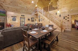 Interior em madeira com uma mesa de jantar posta para quatro pessoas, uma escada e uma sala de estar com sofá e lareira.