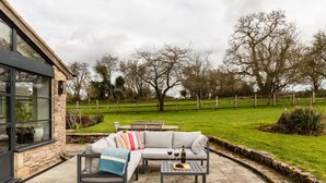 A patio area with a sofa, a table, and a bench. There is a stone wall and a wooden fence in the background. - Lavish Farmhouse With Lake Gardens Pool Table (Radstock)
