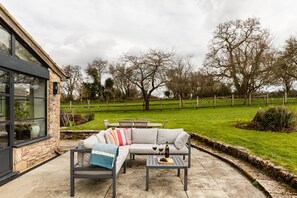 A patio area with a sofa, a table, and a bench. There is a stone wall and a wooden fence in the background. - Lavish Farmhouse With Lake Gardens Pool Table (Radstock)