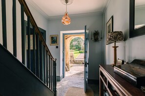 A hallway with a wooden staircase, a door leading outside, and a lamp on a table. - Lavish Farmhouse With Lake Gardens Pool Table (Radstock)