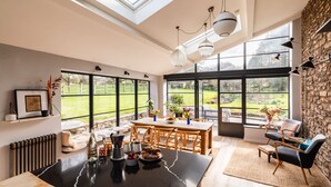 A spacious room with a large dining table, multiple windows offering a view of a garden, and a stone wall. - Lavish Farmhouse With Lake Gardens Pool Table (Radstock)
